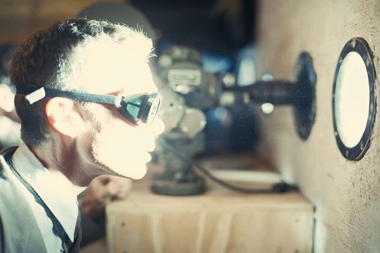 Oppenheimer blickt mit einer aufgesetzten Schweißerbrille durch ein Bullauge.